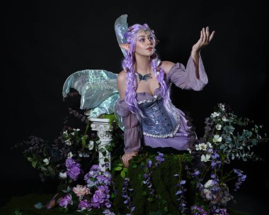 Peri kanatlı ve mor elbiseli fantezi elf kostümü giyen güzel bir kadın modelin tam boy portresi. Oturma pozu, sihirli bir büyü yapar gibi uzanan eller romantik yosunlu büyülü arka plan üzerine.