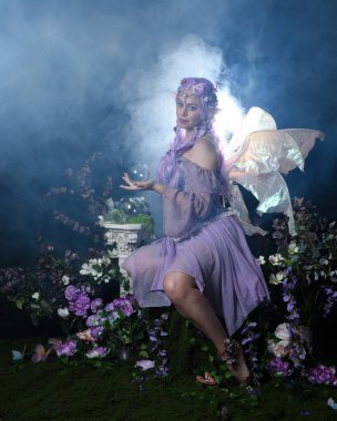 Peri kanatlı ve mor elbiseli fantezi elf kostümü giyen güzel bir kadın model. Oturma pozu kristal küre, sihirli karanlık stüdyo arka planı romantik çiçekler.