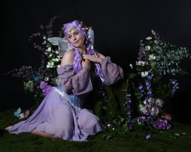 Peri kanatlı ve mor elbiseli fantezi elf kostümü giyen güzel bir kadın model. Oturma pozu, romantik çiçekli sihirli karanlık stüdyo arka planı.
