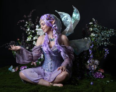 Peri kanatlı ve mor elbiseli fantezi elf kostümü giyen güzel bir kadın model. Oturma pozu, romantik çiçekli sihirli karanlık stüdyo arka planı.
