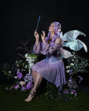 Peri kanatlı ve mor elbiseli fantezi elf kostümü giyen güzel bir kadın model. Oturma pozu sihirli kristal değnek, romantik çiçekli karanlık stüdyo arka planı.