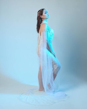 Beyaz inci bir elbise giyen kızıl saçlı bir deniz kızının fantezi stüdyosu portresi, peri masalı kostümü. Akan kumaş ve ruhani stüdyo ışıklarıyla klasik bir duruş sergilediler.