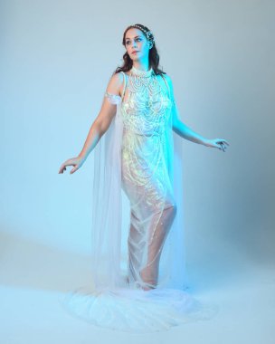 Beyaz inci bir elbise giyen kızıl saçlı bir deniz kızının fantezi stüdyosu portresi, peri masalı kostümü. Akan kumaş ve ruhani stüdyo ışıklarıyla klasik bir duruş sergilediler.