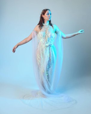 Beyaz inci bir elbise giyen kızıl saçlı bir deniz kızının fantezi stüdyosu portresi, peri masalı kostümü. Akan kumaş ve ruhani stüdyo ışıklarıyla klasik bir duruş sergilediler.