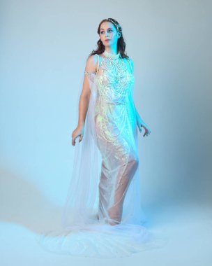 Beyaz inci bir elbise giyen kızıl saçlı bir deniz kızının fantezi stüdyosu portresi, peri masalı kostümü. Akan kumaş ve ruhani stüdyo ışıklarıyla klasik bir duruş sergilediler.