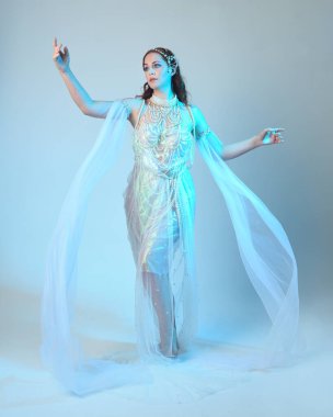 Beyaz inci bir elbise giyen kızıl saçlı bir deniz kızının fantezi stüdyosu portresi, peri masalı kostümü. Akan kumaş ve ruhani stüdyo ışıklarıyla klasik bir duruş sergilediler.