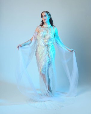 Beyaz inci bir elbise giyen kızıl saçlı bir deniz kızının fantezi stüdyosu portresi, peri masalı kostümü. Akan kumaş ve ruhani stüdyo ışıklarıyla klasik bir duruş sergilediler.