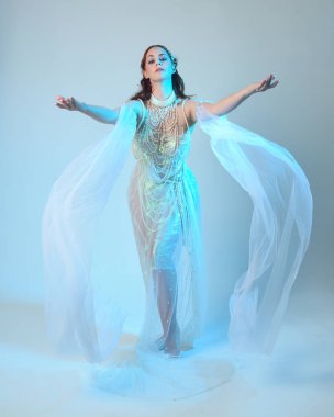 Beyaz inci bir elbise giyen kızıl saçlı bir deniz kızının fantezi stüdyosu portresi, peri masalı kostümü. Akan kumaş ve ruhani stüdyo ışıklarıyla klasik bir duruş sergilediler.