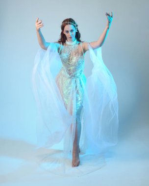 Beyaz inci bir elbise giyen kızıl saçlı bir deniz kızının fantezi stüdyosu portresi, peri masalı kostümü. Akan kumaş ve ruhani stüdyo ışıklarıyla klasik bir duruş sergilediler.