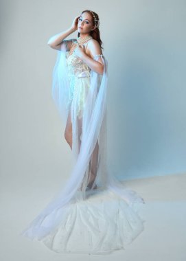 Beyaz inci bir elbise giyen kızıl saçlı bir deniz kızının fantezi stüdyosu portresi, peri masalı kostümü. Akan kumaş ve ruhani stüdyo ışıklarıyla klasik bir duruş sergilediler.