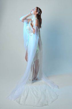 Beyaz inci bir elbise giyen kızıl saçlı bir deniz kızının fantezi stüdyosu portresi, peri masalı kostümü. Akan kumaş ve ruhani stüdyo ışıklarıyla klasik bir duruş sergilediler.