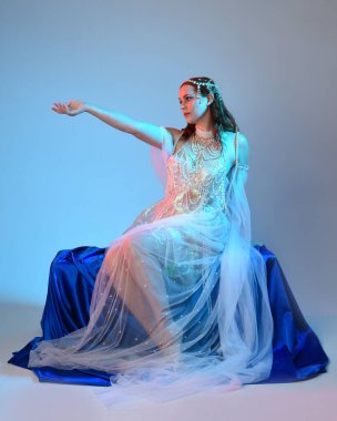 Peri masalı kostümü giymiş kızıl saçlı bir deniz kızı perisinin fantezi stüdyosu portresi. Akan kumaş ve ruhani stüdyo ışıklandırması ile klasik oturma pozisyonu