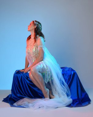 Peri masalı kostümü giymiş kızıl saçlı bir deniz kızı perisinin fantezi stüdyosu portresi. Akan kumaş ve ruhani stüdyo ışıklandırması ile klasik oturma pozisyonu