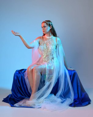 Peri masalı kostümü giymiş kızıl saçlı bir deniz kızı perisinin fantezi stüdyosu portresi. Akan kumaş ve ruhani stüdyo ışıklandırması ile klasik oturma pozisyonu