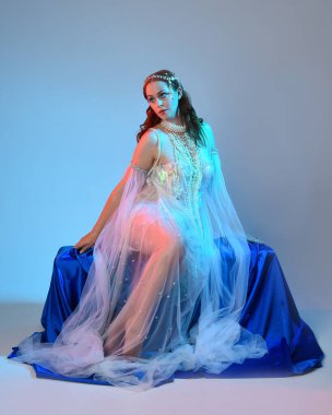 Peri masalı kostümü giymiş kızıl saçlı bir deniz kızı perisinin fantezi stüdyosu portresi. Akan kumaş ve ruhani stüdyo ışıklandırması ile klasik oturma pozisyonu