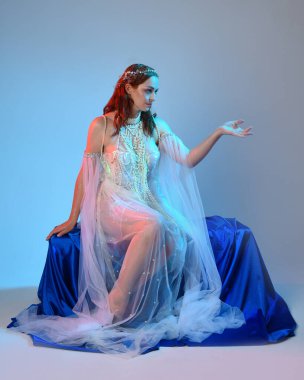 Peri masalı kostümü giymiş kızıl saçlı bir deniz kızı perisinin fantezi stüdyosu portresi. Akan kumaş ve ruhani stüdyo ışıklandırması ile klasik oturma pozisyonu