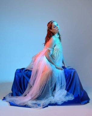 Peri masalı kostümü giymiş kızıl saçlı bir deniz kızı perisinin fantezi stüdyosu portresi. Akan kumaş ve ruhani stüdyo ışıklandırması ile klasik oturma pozisyonu