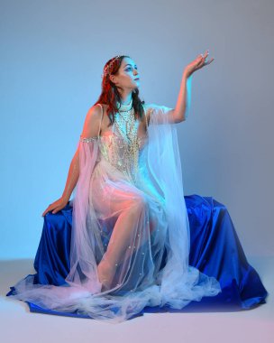 Peri masalı kostümü giymiş kızıl saçlı bir deniz kızı perisinin fantezi stüdyosu portresi. Akan kumaş ve ruhani stüdyo ışıklandırması ile klasik oturma pozisyonu