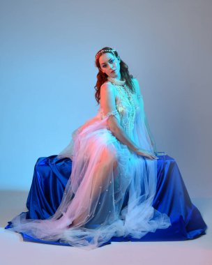 Peri masalı kostümü giymiş kızıl saçlı bir deniz kızı perisinin fantezi stüdyosu portresi. Akan kumaş ve ruhani stüdyo ışıklandırması ile klasik oturma pozisyonu