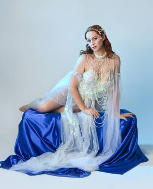 Peri masalı kostümü giymiş kızıl saçlı bir deniz kızı perisinin fantezi stüdyosu portresi. Akan kumaş ve ruhani stüdyo ışıklandırması ile klasik oturma pozisyonu