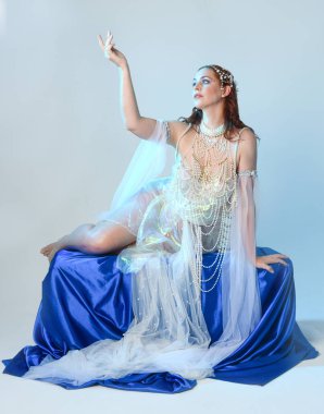 Peri masalı kostümü giymiş kızıl saçlı bir deniz kızı perisinin fantezi stüdyosu portresi. Akan kumaş ve ruhani stüdyo ışıklandırması ile klasik oturma pozisyonu