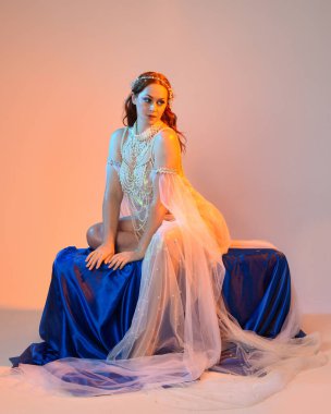 Peri masalı kostümü giymiş kızıl saçlı bir deniz kızı perisinin fantezi stüdyosu portresi. Akan kumaş ve ruhani altın renkli stüdyo ışıklarıyla klasik bir oturma pozisyonu.