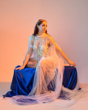 Peri masalı kostümü giymiş kızıl saçlı bir deniz kızı perisinin fantezi stüdyosu portresi. Akan kumaş ve ruhani altın renkli stüdyo ışıklarıyla klasik bir oturma pozisyonu.