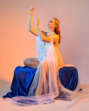 Peri masalı kostümü giymiş kızıl saçlı bir deniz kızı perisinin fantezi stüdyosu portresi. Akan kumaş ve ruhani altın renkli stüdyo ışıklarıyla klasik bir oturma pozisyonu.