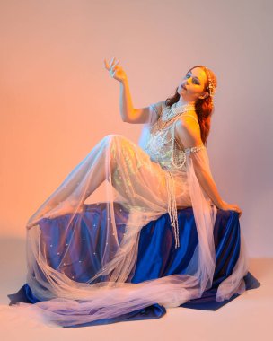 Peri masalı kostümü giymiş kızıl saçlı bir deniz kızı perisinin fantezi stüdyosu portresi. Akan kumaş ve ruhani altın renkli stüdyo ışıklarıyla klasik bir oturma pozisyonu.