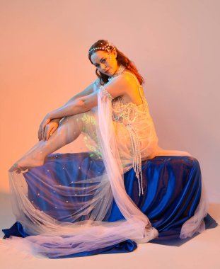 Peri masalı kostümü giymiş kızıl saçlı bir deniz kızı perisinin fantezi stüdyosu portresi. Akan kumaş ve ruhani altın renkli stüdyo ışıklarıyla klasik bir oturma pozisyonu.