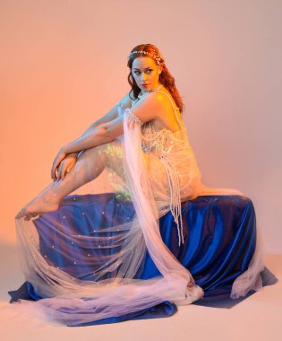 Peri masalı kostümü giymiş kızıl saçlı bir deniz kızı perisinin fantezi stüdyosu portresi. Akan kumaş ve ruhani altın renkli stüdyo ışıklarıyla klasik bir oturma pozisyonu.