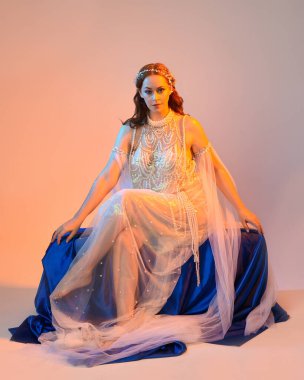 Peri masalı kostümü giymiş kızıl saçlı bir deniz kızı perisinin fantezi stüdyosu portresi. Akan kumaş ve ruhani altın renkli stüdyo ışıklarıyla klasik bir oturma pozisyonu.