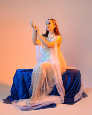 Peri masalı kostümü giymiş kızıl saçlı bir deniz kızı perisinin fantezi stüdyosu portresi. Akan kumaş ve ruhani altın renkli stüdyo ışıklarıyla klasik bir oturma pozisyonu.