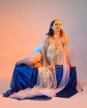 Peri masalı kostümü giymiş kızıl saçlı bir deniz kızı perisinin fantezi stüdyosu portresi. Akan kumaş ve ruhani altın renkli stüdyo ışıklarıyla klasik bir oturma pozisyonu.