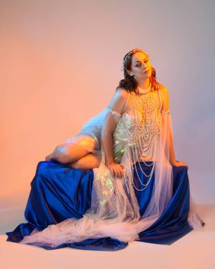 Peri masalı kostümü giymiş kızıl saçlı bir deniz kızı perisinin fantezi stüdyosu portresi. Akan kumaş ve ruhani altın renkli stüdyo ışıklarıyla klasik bir oturma pozisyonu.