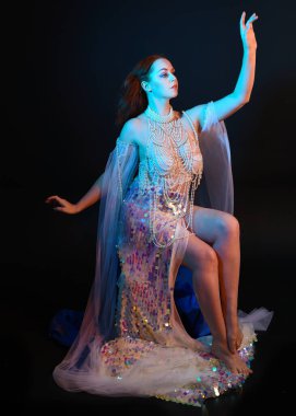 Peri masalı kostümü giymiş kızıl saçlı bir deniz kızı perisinin fantezi stüdyosu portresi. Akan kumaş ve ruhsal kasvetli stüdyo ışıklandırmasıyla klasik bir oturma pozisyonu.