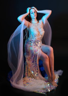 Peri masalı kostümü giymiş kızıl saçlı bir deniz kızı perisinin fantezi stüdyosu portresi. Akan kumaş ve ruhsal kasvetli stüdyo ışıklandırmasıyla klasik bir oturma pozisyonu.