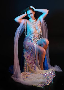 Peri masalı kostümü giymiş kızıl saçlı bir deniz kızı perisinin fantezi stüdyosu portresi. Akan kumaş ve ruhsal kasvetli stüdyo ışıklandırmasıyla klasik bir oturma pozisyonu.