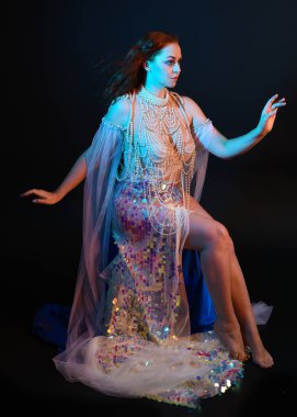 Peri masalı kostümü giymiş kızıl saçlı bir deniz kızı perisinin fantezi stüdyosu portresi. Akan kumaş ve ruhsal kasvetli stüdyo ışıklandırmasıyla klasik bir oturma pozisyonu.