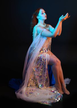 Peri masalı kostümü giymiş kızıl saçlı bir deniz kızı perisinin fantezi stüdyosu portresi. Akan kumaş ve ruhsal kasvetli stüdyo ışıklandırmasıyla klasik bir oturma pozisyonu.