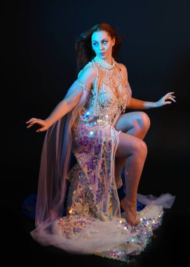 Peri masalı kostümü giymiş kızıl saçlı bir deniz kızı perisinin fantezi stüdyosu portresi. Akan kumaş ve ruhsal kasvetli stüdyo ışıklandırmasıyla klasik bir oturma pozisyonu.