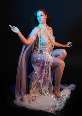 Peri masalı kostümü giymiş kızıl saçlı bir deniz kızı perisinin fantezi stüdyosu portresi. Akan kumaş ve ruhsal kasvetli stüdyo ışıklandırmasıyla klasik bir oturma pozisyonu.