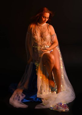 Peri masalı kostümü giymiş kızıl saçlı bir deniz kızı perisinin fantezi stüdyosu portresi. Akan kumaş ve ruhsal kasvetli stüdyo ışıklandırmasıyla klasik bir oturma pozisyonu.