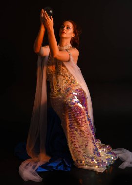 Peri masalı kostümü giymiş kızıl saçlı bir deniz kızı perisinin fantezi stüdyosu portresi. Akan kumaş ve ruhsal kasvetli stüdyo ışıklandırmasıyla klasik bir oturma pozisyonu.