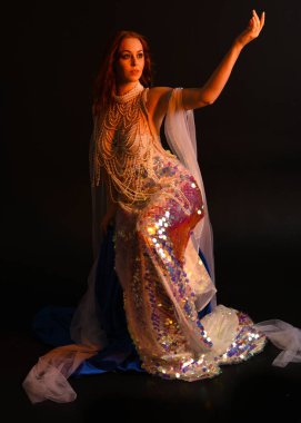 Peri masalı kostümü giymiş kızıl saçlı bir deniz kızı perisinin fantezi stüdyosu portresi. Akan kumaş ve ruhsal kasvetli stüdyo ışıklandırmasıyla klasik bir oturma pozisyonu.