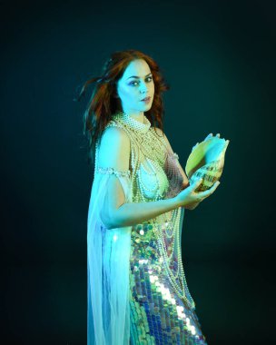 Peri masalı kostümü giymiş kızıl saçlı bir deniz kızı perisinin fantezi stüdyosu portresi. Klasik poz, deniz kabuğunu havada süzülen kumaş ve ruhsal kasvetli stüdyo ışıklandırmasıyla tutuyordu.