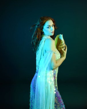 Peri masalı kostümü giymiş kızıl saçlı bir deniz kızı perisinin fantezi stüdyosu portresi. Klasik poz, deniz kabuğunu havada süzülen kumaş ve ruhsal kasvetli stüdyo ışıklandırmasıyla tutuyordu.