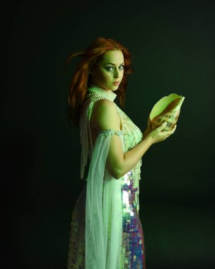 Peri masalı kostümü giymiş kızıl saçlı bir deniz kızı perisinin fantezi stüdyosu portresi. Klasik poz, deniz kabuğunu havada süzülen kumaş ve ruhsal kasvetli stüdyo ışıklandırmasıyla tutuyordu.