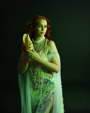 Peri masalı kostümü giymiş kızıl saçlı bir deniz kızı perisinin fantezi stüdyosu portresi. Klasik poz, deniz kabuğunu havada süzülen kumaş ve ruhsal kasvetli stüdyo ışıklandırmasıyla tutuyordu.