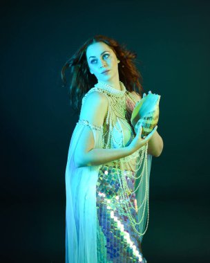 Peri masalı kostümü giymiş kızıl saçlı bir deniz kızı perisinin fantezi stüdyosu portresi. Klasik poz, deniz kabuğunu havada süzülen kumaş ve ruhsal kasvetli stüdyo ışıklandırmasıyla tutuyordu.